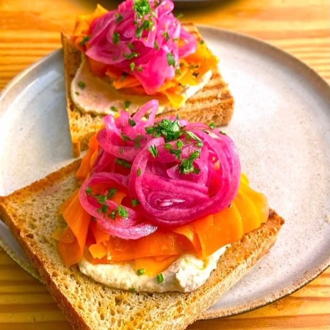 Duas tostas com fitas de cenoura, cebola roxa e ervas sobre creme branco, num prato claro