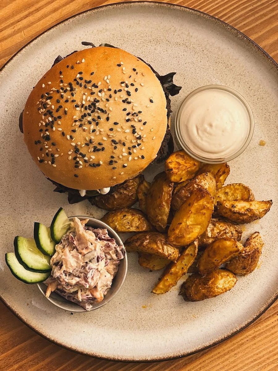 Hambúrguer com sésamo, batatas em gomos, coleslaw, pepino e molho branco num prato claro