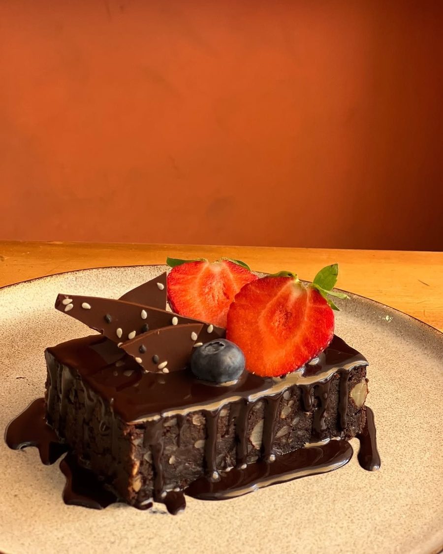 Brownie com molho de chocolate, morangos e um mirtilo, diante de uma parede cor de terracota