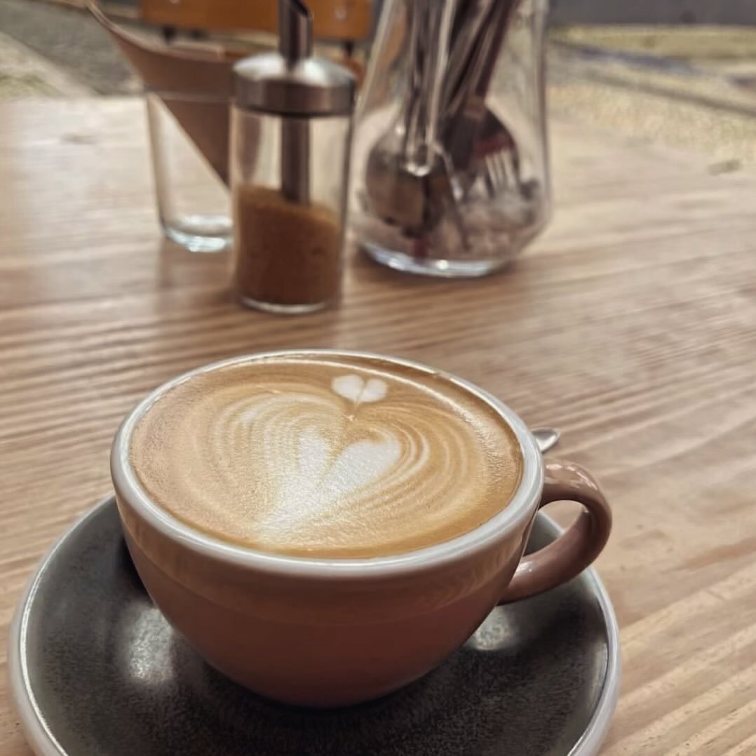 Cappuccino com desenho no leite numa mesa de madeira, com açucareiro e talheres ao fundo