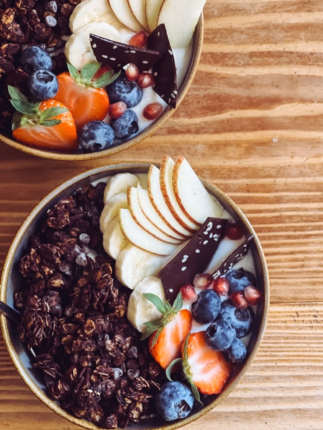 Duas taças de granola escura com morangos, mirtilos, banana e lascas de chocolate sobre uma mesa de madeira